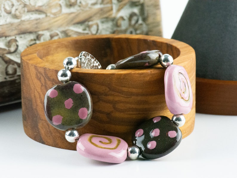 May include: A bracelet made of brown and pink ceramic beads with silver accents. The beads are patterned with polka dots and swirls.