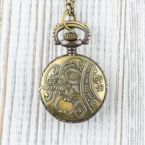 Puede incluir: Un reloj de bolsillo dorado con un dise&ntilde;o floral en la esfera. El reloj est&aacute; colgado de una cadena sobre un fondo de madera blanca.
