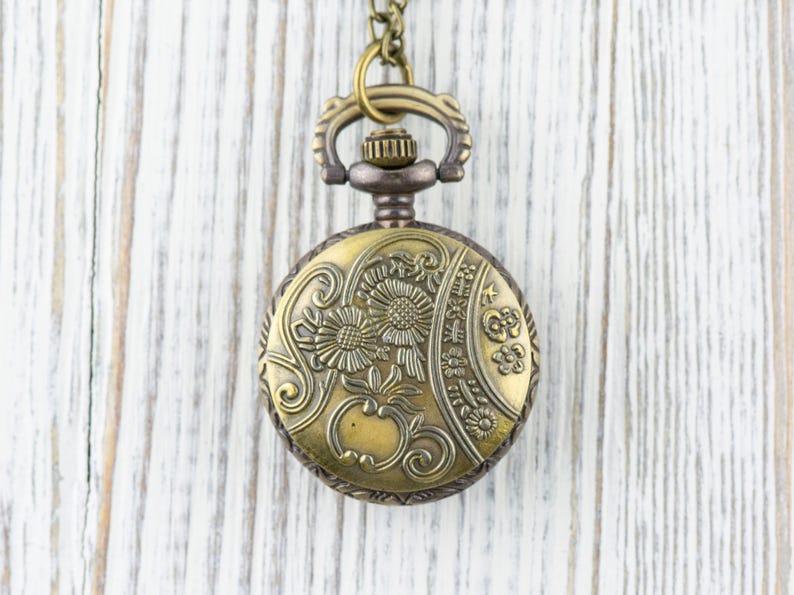 Puede incluir: Un reloj de bolsillo dorado con un dise&ntilde;o floral en la esfera. El reloj est&aacute; colgado de una cadena sobre un fondo de madera blanca.