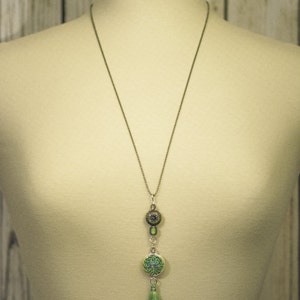 May include: A silver chain necklace with a green and silver pendant featuring a tree design. The pendant is attached to a green tassel.