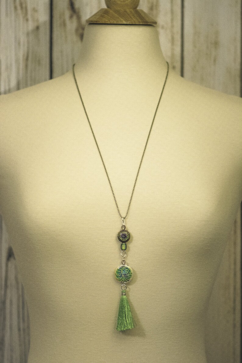 May include: A silver chain necklace with a green and silver pendant featuring a tree design. The pendant is attached to a green tassel.