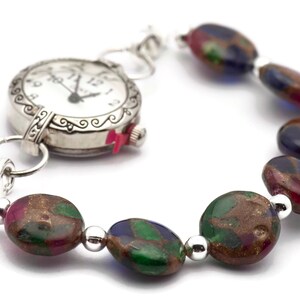 May include: A silver watch with a white face and black hands is attached to a bracelet made of colorful, oval-shaped beads. The beads are a mix of green, blue, red, and gold.
