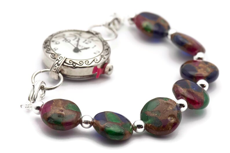 May include: A silver watch with a white face and black hands is attached to a bracelet made of colorful, oval-shaped beads. The beads are a mix of green, blue, red, and gold.
