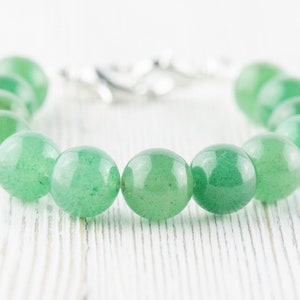 May include: A green gemstone bracelet with a silver clasp. The bracelet is made of round beads and is lying on a white wooden surface.