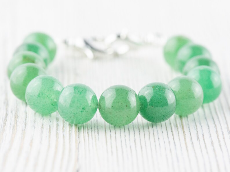 May include: A green gemstone bracelet with a silver clasp. The bracelet is made of round beads and is lying on a white wooden surface.