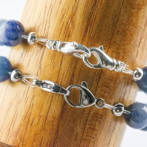 May include: Two blue gemstone bracelets with silver clasps and silver beads. The bracelets are stacked on top of each other on a wooden surface.