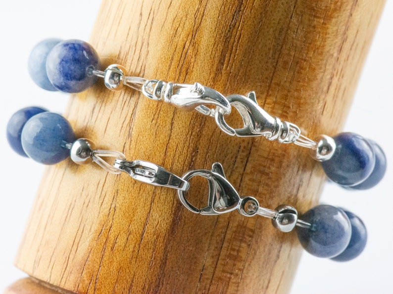 May include: Two blue gemstone bracelets with silver clasps and silver beads. The bracelets are stacked on top of each other on a wooden surface.