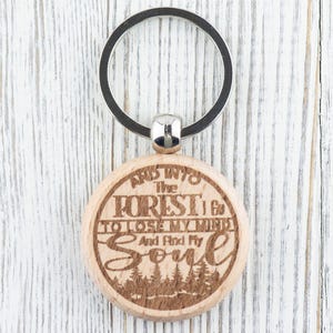 Op de afbeelding: Houten sleutelhanger met een rond, gegraveerd label met de tekst "And into the forest I go to lose my mind and find my soul". Het label is bevestigd aan een zilveren sleutelring.