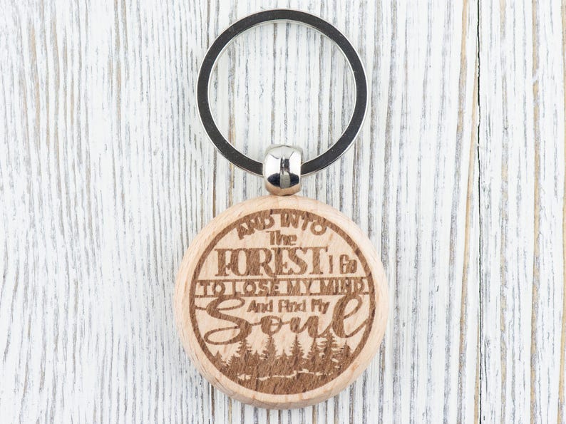Op de afbeelding: Houten sleutelhanger met een rond, gegraveerd label met de tekst "And into the forest I go to lose my mind and find my soul". Het label is bevestigd aan een zilveren sleutelring.