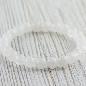 May include: A white gemstone bracelet made of smooth, oval-shaped beads. The bracelet is lying on a white wooden surface.