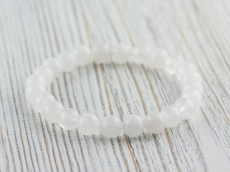 May include: A white gemstone bracelet made of smooth, oval-shaped beads. The bracelet is lying on a white wooden surface.
