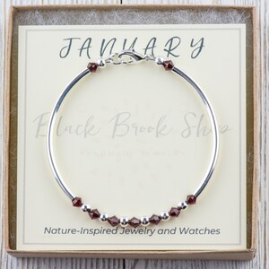 May include: A silver bangle bracelet with a red gemstone charm. The bracelet is in a box with the text "JANUARY" and "Nature-Inspired Jewelry and Watches".