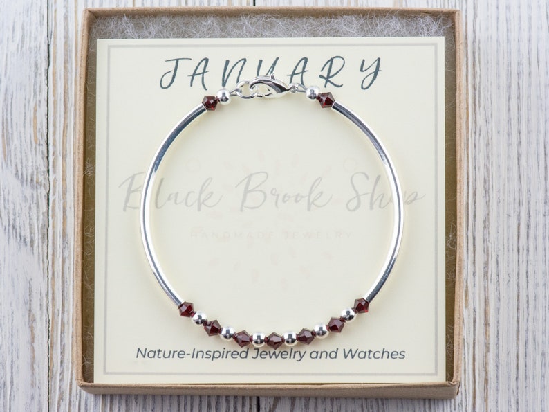 May include: A silver bangle bracelet with a red gemstone charm. The bracelet is in a box with the text "JANUARY" and "Nature-Inspired Jewelry and Watches".
