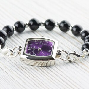 May include: A silver watch with a square face and a purple dial. The watch is attached to a black beaded bracelet.