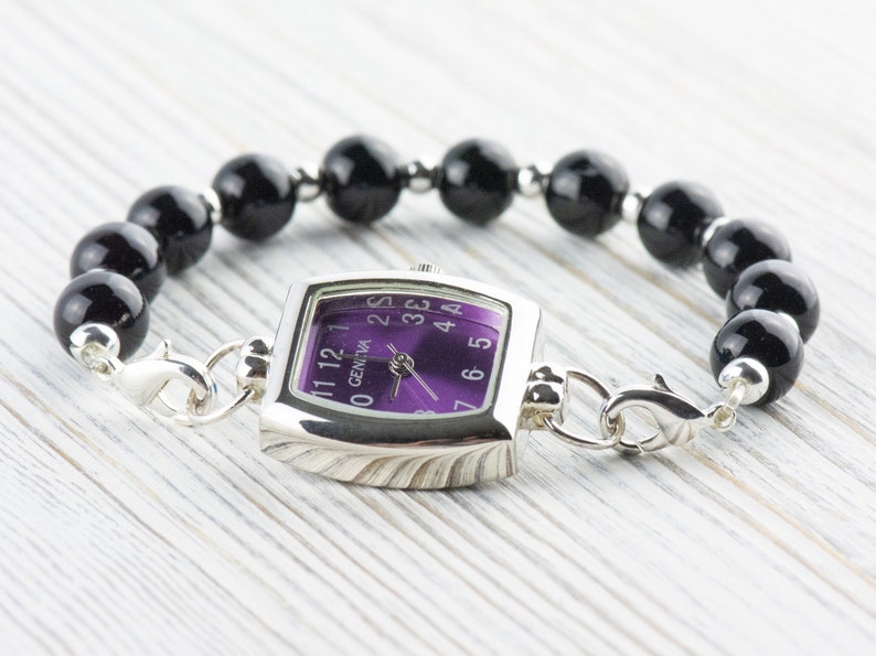 May include: A silver watch with a square face and a purple dial. The watch is attached to a black beaded bracelet.