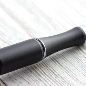 May include: A close-up of a black bottle stopper with a silver accent ring and a silver tip. The stopper has a matte finish and a rounded, ergonomic design. The background is a light-colored, wooden surface.