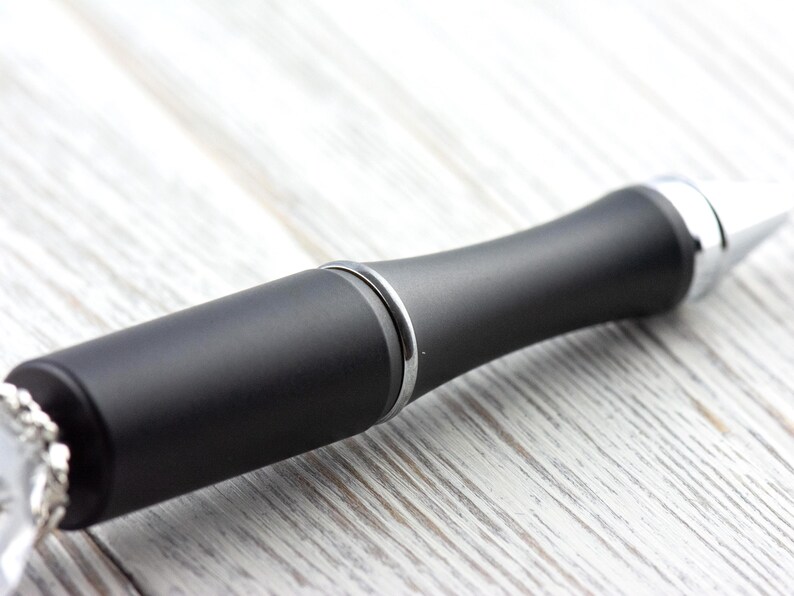 May include: A close-up of a black bottle stopper with a silver accent ring and a silver tip. The stopper has a matte finish and a rounded, ergonomic design. The background is a light-colored, wooden surface.