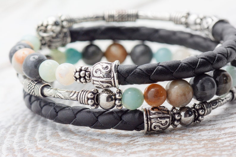 May include: A triple-wrap leather bracelet with silver beads and charms. The bracelet features a braided black leather band with silver beads and charms, including a decorative silver bead with an inscription.