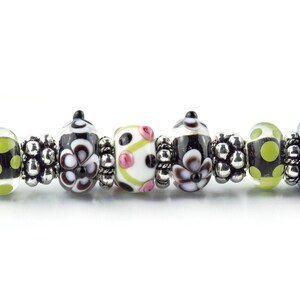 Op de afbeelding: Close-up van een kralenarmband met diverse glazen kralen. De kralen hebben zwart-witte bloempatronen, stippen en roze accenten. Zilverkleurige afstandhouders scheiden de kralen. De armband heeft zwarte uiteinden.