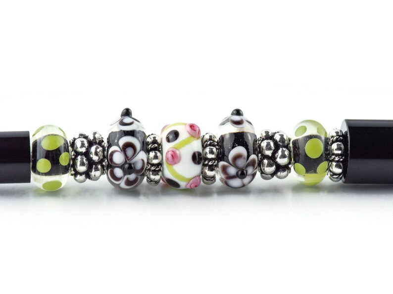 Op de afbeelding: Close-up van een kralenarmband met diverse glazen kralen. De kralen hebben zwart-witte bloempatronen, stippen en roze accenten. Zilverkleurige afstandhouders scheiden de kralen. De armband heeft zwarte uiteinden.