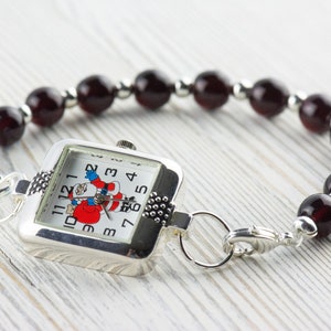 Pu&ograve; includere: Un orologio d'argento con un quadrante quadrato e un design di Babbo Natale rosso e bianco. L'orologio &egrave; attaccato a un braccialetto fatto di perline rosso scuro.
