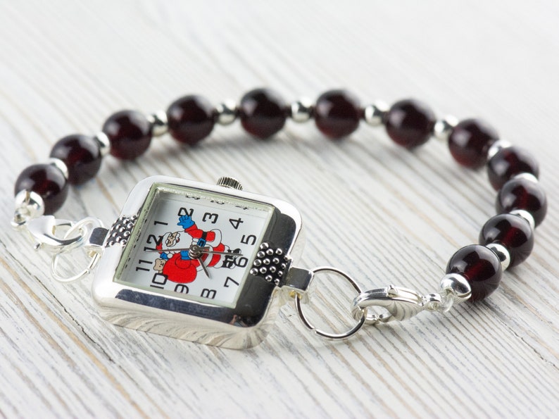 Pu&ograve; includere: Un orologio d'argento con un quadrante quadrato e un design di Babbo Natale rosso e bianco. L'orologio &egrave; attaccato a un braccialetto fatto di perline rosso scuro.