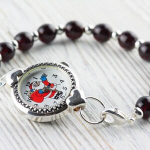 Peut inclure: Une montre argent&eacute;e avec un visage de P&egrave;re No&euml;l sur le cadran, attach&eacute;e &agrave; un bracelet compos&eacute; de perles rouge fonc&eacute;.