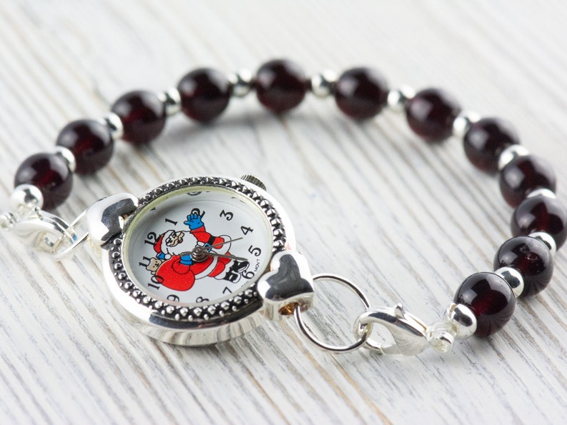 Peut inclure: Une montre argent&eacute;e avec un visage de P&egrave;re No&euml;l sur le cadran, attach&eacute;e &agrave; un bracelet compos&eacute; de perles rouge fonc&eacute;.