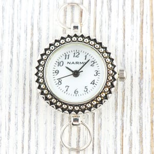 K&ouml;nnte beinhalten: Eine silberfarbene Taschenuhr mit wei&szlig;em Zifferblatt und schwarzen Zeigern. Die Uhr hat eine dekorative L&uuml;nette mit Perlenrand und Ringe zur Befestigung. Das Zifferblatt zeigt den Markennamen "NARM".