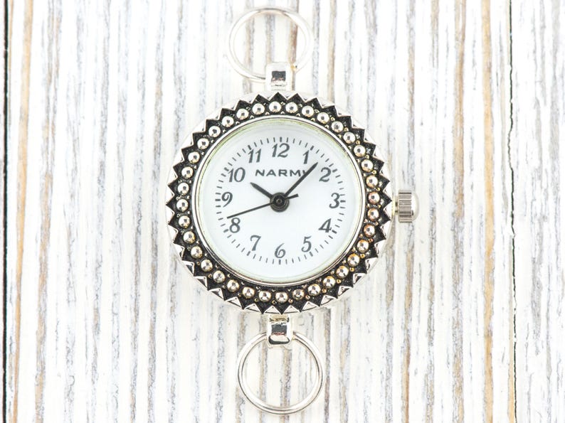 K&ouml;nnte beinhalten: Eine silberfarbene Taschenuhr mit wei&szlig;em Zifferblatt und schwarzen Zeigern. Die Uhr hat eine dekorative L&uuml;nette mit Perlenrand und Ringe zur Befestigung. Das Zifferblatt zeigt den Markennamen "NARM".