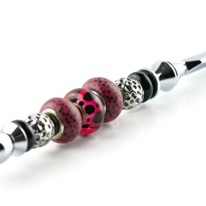 May include: A silver-toned metal wand with decorative beads. The beads include a mix of textures and colors, featuring red and black spotted beads, silver filigree beads, and black beads. The wand has a rounded tip and a decorative end.