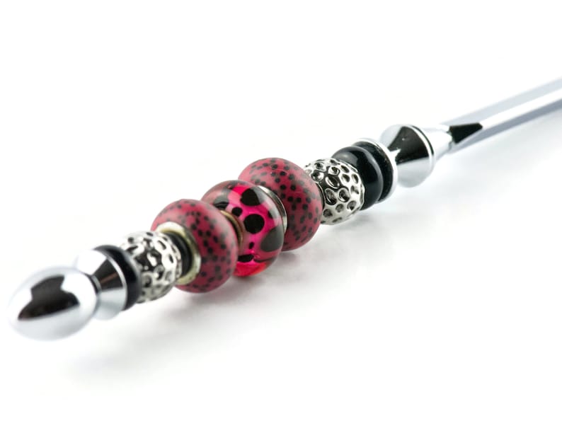 May include: A silver-toned metal wand with decorative beads. The beads include a mix of textures and colors, featuring red and black spotted beads, silver filigree beads, and black beads. The wand has a rounded tip and a decorative end.
