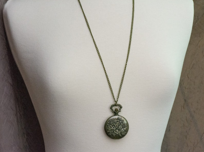 May include: A bronze-colored pocket watch necklace with a floral design on the face. The chain is a simple, linked chain.