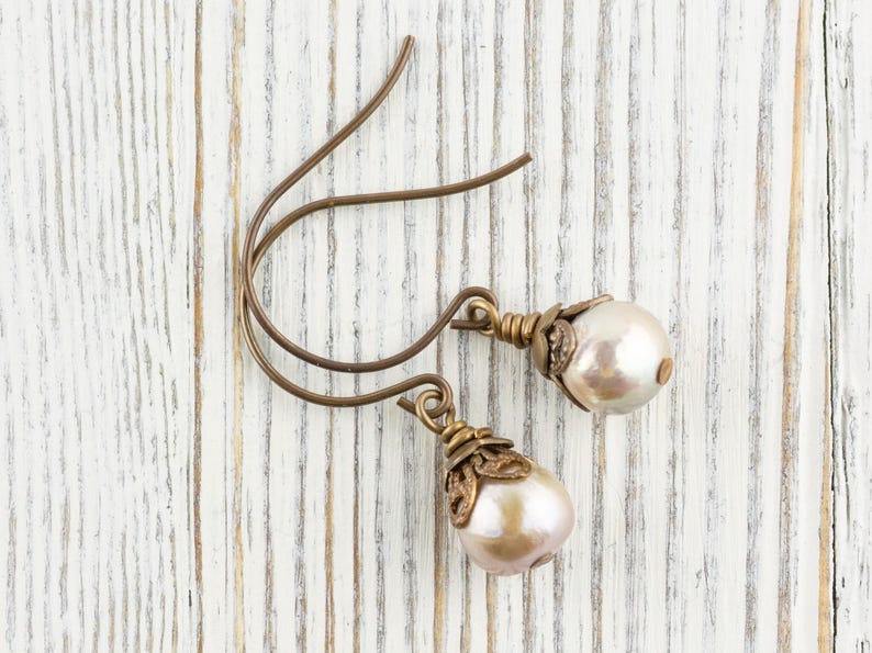 May include: A pair of drop earrings with pearlescent, teardrop-shaped pearls. The earrings have bronze-colored, decorative caps and ear wires. The earrings are displayed on a white, weathered wooden surface.