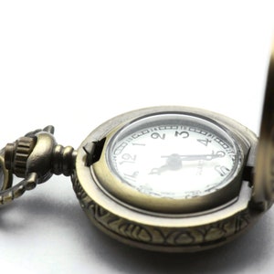 Puede incluir: Un reloj de bolsillo de estilo vintage con un acabado dorado y un dise&ntilde;o ornamentado. El reloj tiene una esfera blanca con marcadores de hora en n&uacute;meros romanos negros y una aguja de segundos negra. El reloj est&aacute; abierto y la cadena es visible.