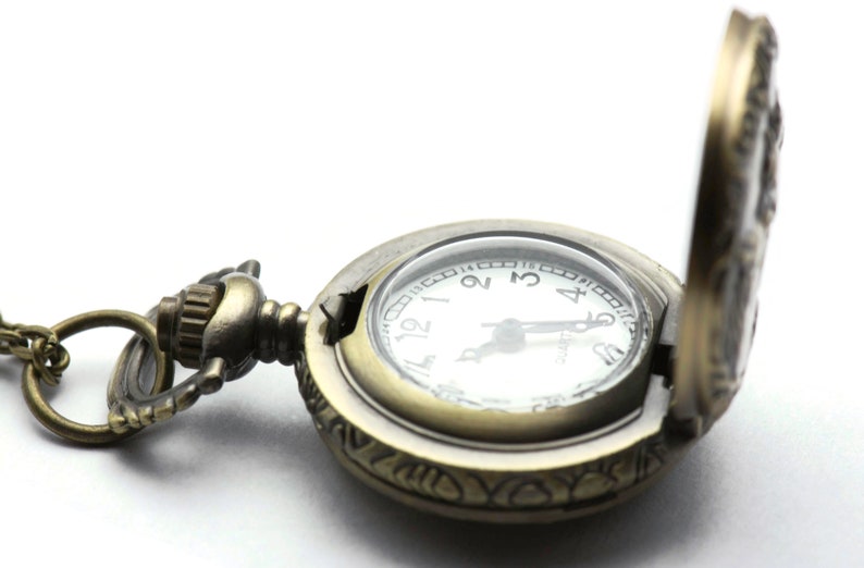 Puede incluir: Un reloj de bolsillo de estilo vintage con un acabado dorado y un dise&ntilde;o ornamentado. El reloj tiene una esfera blanca con marcadores de hora en n&uacute;meros romanos negros y una aguja de segundos negra. El reloj est&aacute; abierto y la cadena es visible.
