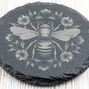 May include: Round, dark gray slate coaster with a detailed etched design of a bee surrounded by flowers and leaves. The bee has outstretched wings and is centered on the coaster. The floral design is symmetrical, with flowers and leaves around the bee.