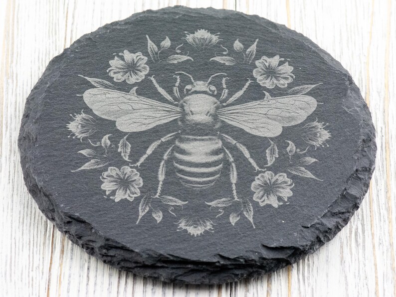 May include: Round, dark gray slate coaster with a detailed etched design of a bee surrounded by flowers and leaves. The bee has outstretched wings and is centered on the coaster. The floral design is symmetrical, with flowers and leaves around the bee.