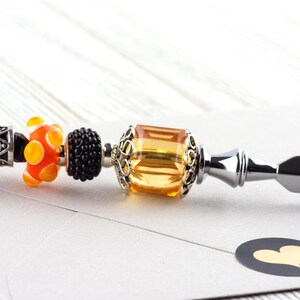 May include: A decorative letter opener with a silver handle and a series of colorful beads. The beads include orange and yellow glass beads, a black beaded bead, and a large amber-colored bead. A black sticker with a gold heart is in the foreground.