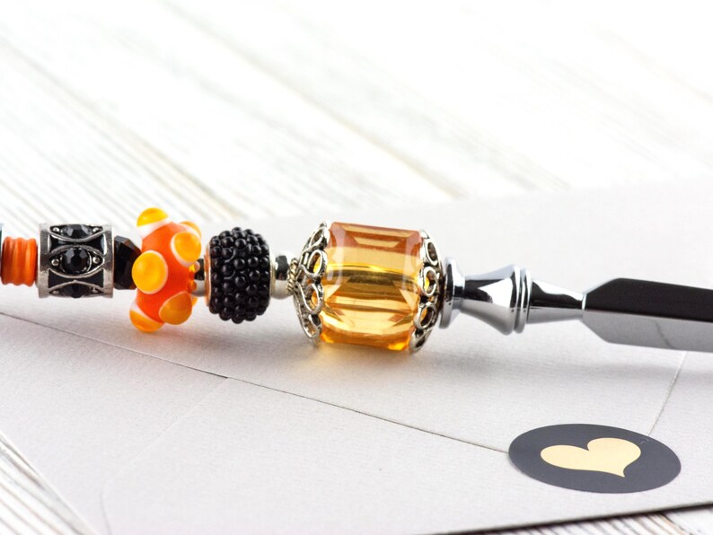 May include: A decorative letter opener with a silver handle and a series of colorful beads. The beads include orange and yellow glass beads, a black beaded bead, and a large amber-colored bead. A black sticker with a gold heart is in the foreground.