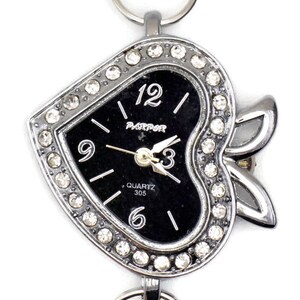 Könnte beinhalten: Eine silberne herzförmige Uhr mit schwarzem Zifferblatt und Strasssteinen. Die Uhr hat ein Quarzwerk und ist mit "QUARTZ 305" beschriftet.