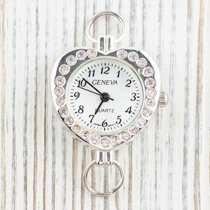 May include: Silver heart-shaped watch with a white face and rhinestone accents. The watch face reads "GENEVA QUARTZ" and has black hands and black numbers.