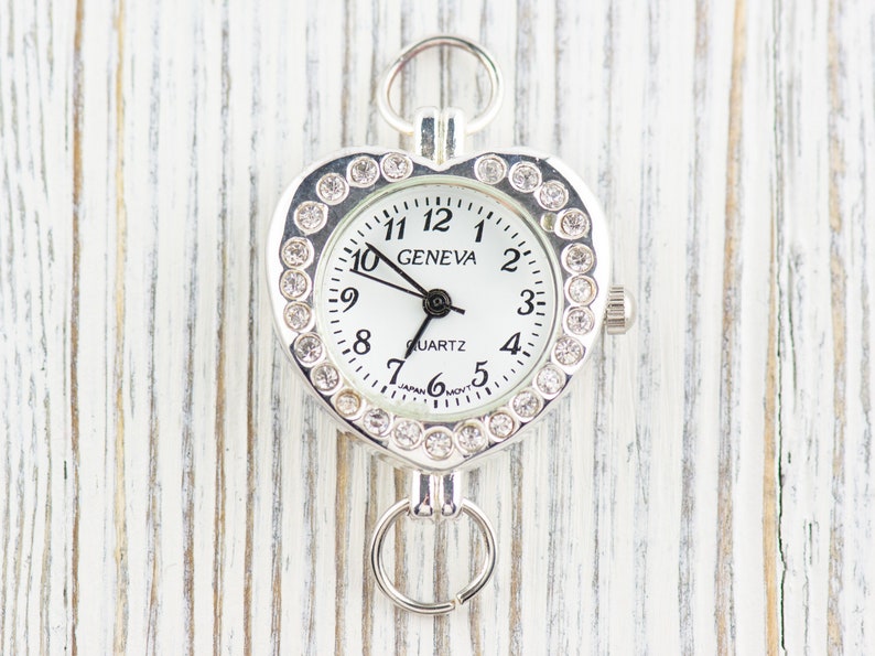 Pu&ograve; includere: Orologio a forma di cuore argentato con quadrante bianco e dettagli in strass. Il quadrante dell'orologio riporta la scritta "GENEVA QUARTZ" e ha lancette nere e numeri neri.