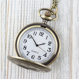 Pode incluir: Um rel&oacute;gio de bolso dourado com um mostrador branco e ponteiros pretos. O rel&oacute;gio tem uma corrente presa a ele e est&aacute; colocado sobre um fundo de madeira branca. O mostrador do rel&oacute;gio tem marcadores de hora em algarismos romanos pretos e a palavra "QUARTZ" impressa nele.