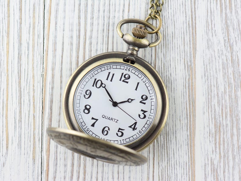 Pode incluir: Um rel&oacute;gio de bolso dourado com um mostrador branco e ponteiros pretos. O rel&oacute;gio tem uma corrente presa a ele e est&aacute; colocado sobre um fundo de madeira branca. O mostrador do rel&oacute;gio tem marcadores de hora em algarismos romanos pretos e a palavra "QUARTZ" impressa nele.