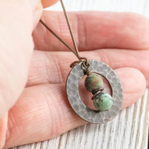 Puede incluir: Un collar con colgante hecho a mano con un c&iacute;rculo de metal plateado martillado y dos cuentas de piedra verde y marr&oacute;n. El collar tiene una est&eacute;tica r&uacute;stica y terrosa. El colgante est&aacute; sujeto por una mano. Ideal para el uso diario.