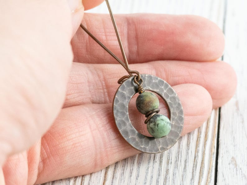 Puede incluir: Un collar con colgante hecho a mano con un c&iacute;rculo de metal plateado martillado y dos cuentas de piedra verde y marr&oacute;n. El collar tiene una est&eacute;tica r&uacute;stica y terrosa. El colgante est&aacute; sujeto por una mano. Ideal para el uso diario.