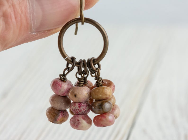 Op de afbeelding: Een handgemaakte oorbel met een bronzen ring, met meerdere strengen kleine, afgeronde kralen. De kralen zijn een mix van roze, bruine en bruine tinten, wat een natuurlijke uitstraling cre&euml;ert. De oorbel wordt door een hand omhoog gehouden.