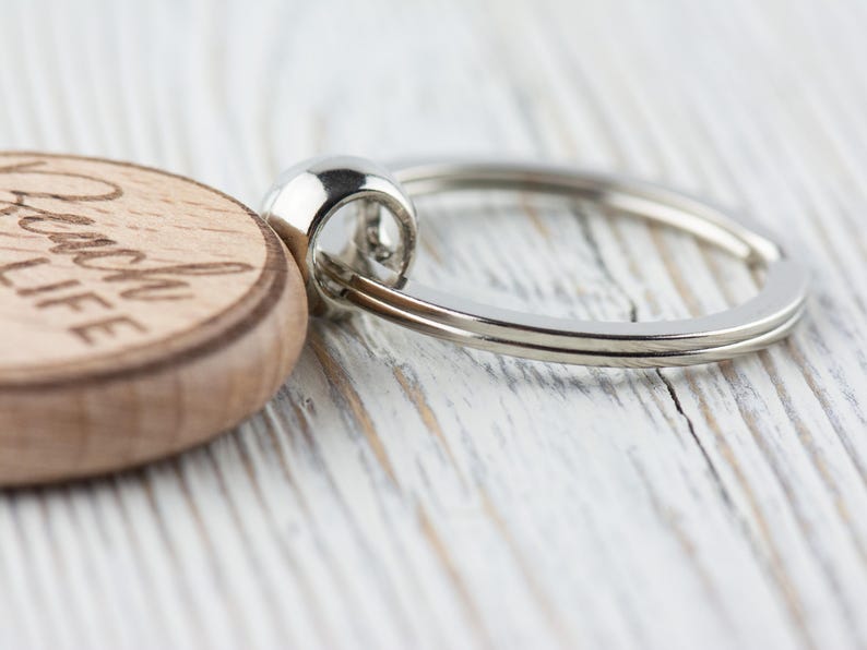 K&ouml;nnte beinhalten: Nahaufnahme eines Holzschl&uuml;sselanh&auml;ngers mit den Worten "Beach Life" eingraviert. Der Schl&uuml;sselanh&auml;nger hat einen silbernen Metallring. Die Holzscheibe ist hellbraun und der Metallring gl&auml;nzend silberfarben.