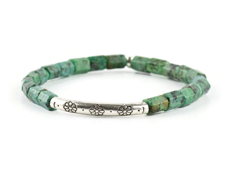 African Turquoise Stretch Bracelet on white background. Beads are bluish green with black and brown speckle. Silver pewter tube focal bead is adorned with floral etchings.